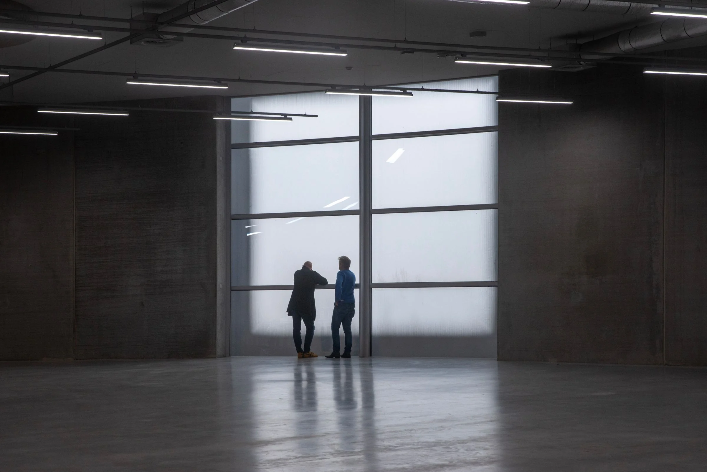 Weite, minimalistische Halle mit hellem Fenster, zwei kleine Figuren in der Ferne
